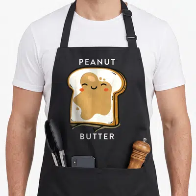 Jelly butter apron - Cartoon toast with tan peanut butter and white PEANUT BUTTER text labels — shown on a black cotton canvas apron against a white background