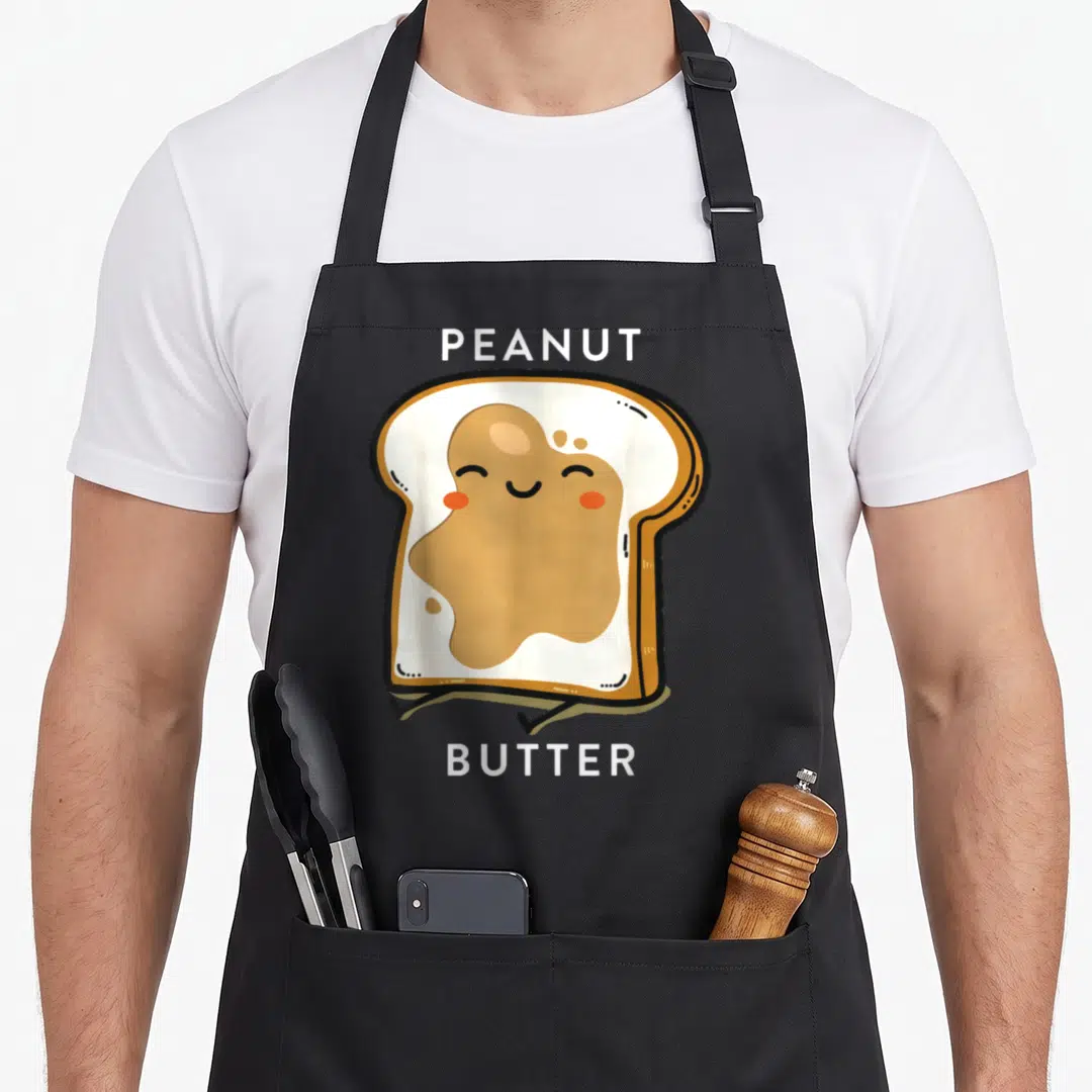 Jelly butter apron - Cartoon toast with tan peanut butter and white PEANUT BUTTER text labels — shown on a black cotton canvas apron against a white background