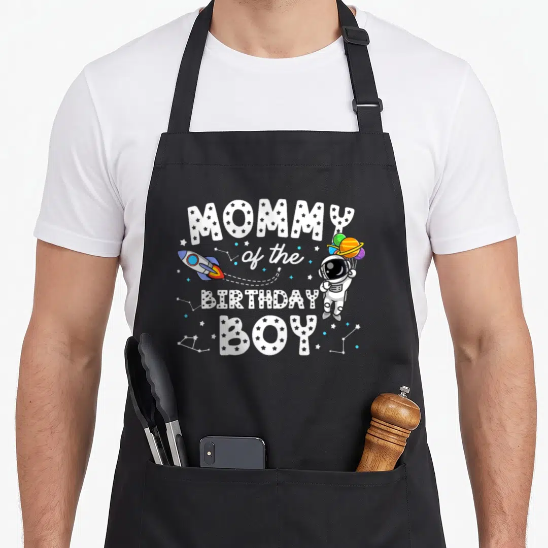 Retro astronaut birthday apron - MOMMY of the BIRTHDAY BOY text with cartoon astronaut and rocket — shown on a black cotton canvas apron against a white background