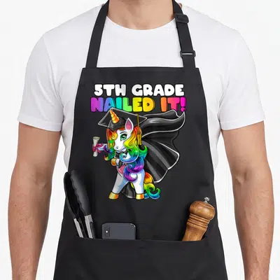Flossing 5th unicorn apron - Cartoon unicorn in cap and gown holding diploma under 5th Grade Nailed It text — shown on a black cotton canvas apron against a white background