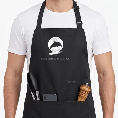 Funny thank fish apron - White distressed dolphin jumping before sun with text So long and thanks for all the fish — shown on a black cotton canvas apron against a white background