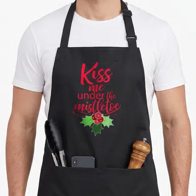 Kiss mistletoe christmas apron - Red script text Kiss me under the mistletoe above green leaves and red berries — shown on a black cotton canvas apron against a white background