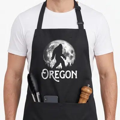Oregon tree apron - Bigfoot silhouette walking before full moon and pine trees above OREGON text — shown on a black cotton canvas apron against a white background