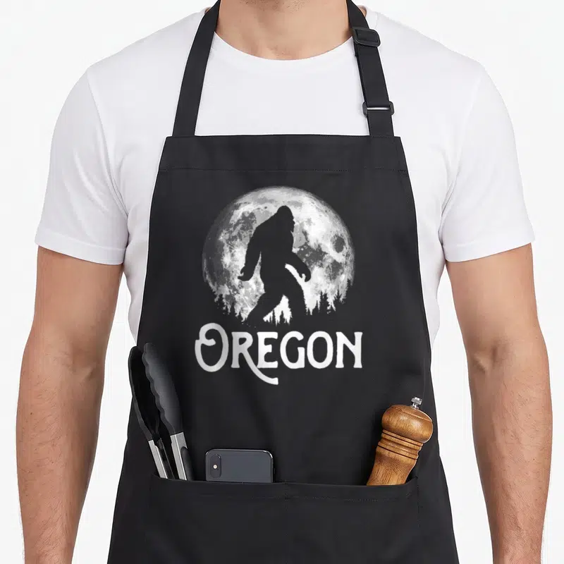Oregon tree apron - Bigfoot silhouette walking before full moon and pine trees above OREGON text — shown on a black cotton canvas apron against a white background