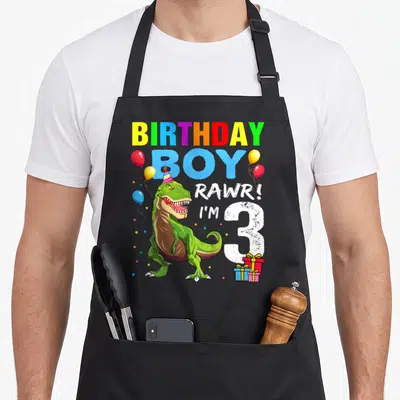 Birthday t rex apron - Green T-Rex wearing party hat with rainbow BIRTHDAY BOY text and number 3 — shown on a black cotton canvas apron against a white background
