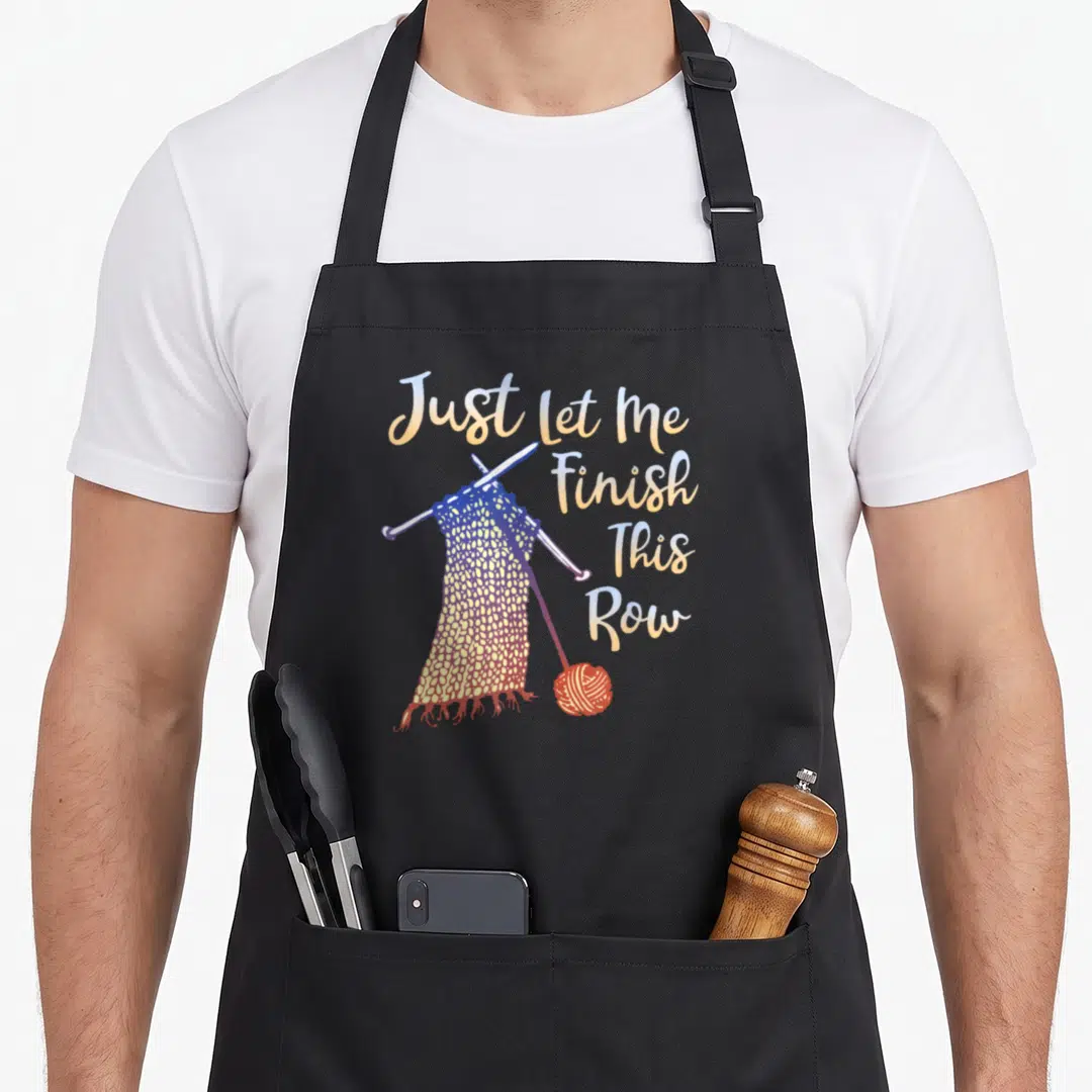 Finish knitting apron - Just Let Me Finish This Row text with knitting needles and yarn ball in yellow blue purple — shown on a black cotton canvas apron against a white background