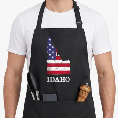 Idaho 4th of july apron - Distressed Idaho state map filled with red white blue flag pattern and white IDAHO text below — shown on a black cotton canvas apron against a white background