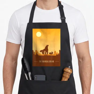 Desert star apron - Brown and orange minimalist silhouette of a person and pod under a yellow sun with centered text — shown on a black cotton canvas apron against a white background