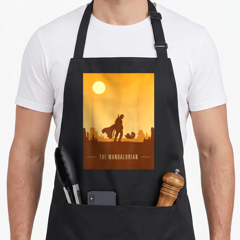 Desert star apron - Brown and orange minimalist silhouette of a person and pod under a yellow sun with centered text — shown on a black cotton canvas apron against a white background