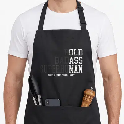 Gag husband apron - Stacked bold bad ass superhuman text in grey and white distressed font on dark background — shown on a black cotton canvas apron against a white background
