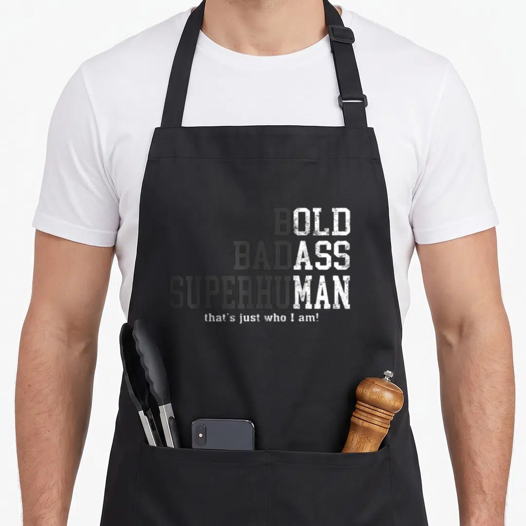 Gag husband apron - Stacked bold bad ass superhuman text in grey and white distressed font on dark background — shown on a black cotton canvas apron against a white background