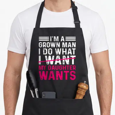 Funny grown father daughter aprons - Stacked white and pink text with pink strikethrough reading I am a grown man my — shown on a black cotton canvas apron against a white background