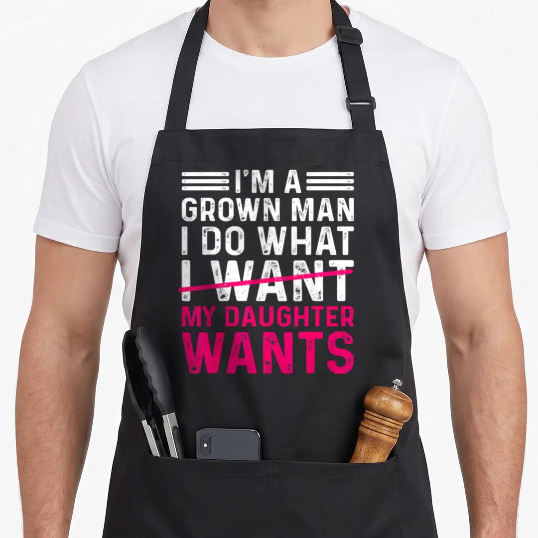 Funny grown father daughter aprons - Stacked white and pink text with pink strikethrough reading I am a grown man my — shown on a black cotton canvas apron against a white background