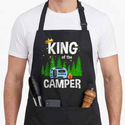 King trailer apron - King of the Camper text with blue trailer, green trees, gold crown, and yellow stars — shown on a black cotton canvas apron against a white background