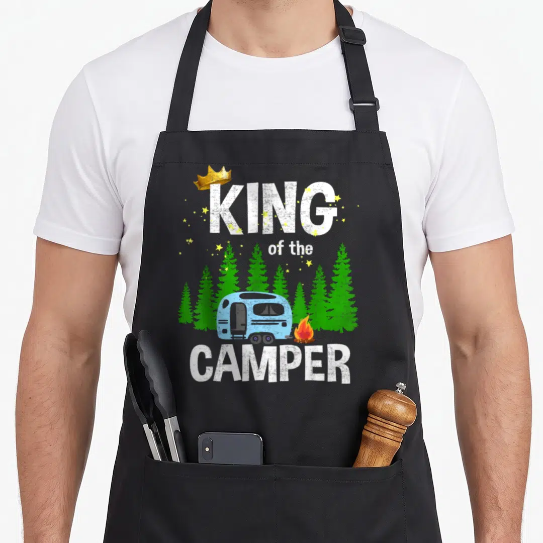 King trailer apron - King of the Camper text with blue trailer, green trees, gold crown, and yellow stars — shown on a black cotton canvas apron against a white background