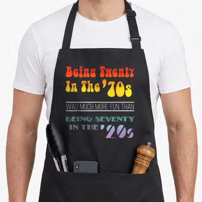 Twenty 70s apron - Retro bubble and striped typography in orange yellow blue and purple with white separating lines — shown on a black cotton canvas apron against a white background