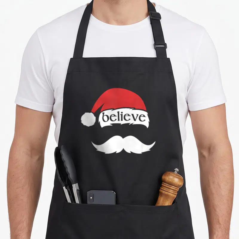 Mustache santa aprons - Red Santa hat with believe text and white mustache graphic — shown on a black cotton canvas apron against a white background