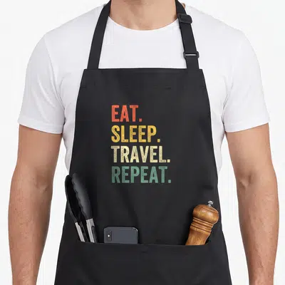 Eat travel aprons - Stacked EAT SLEEP TRAVEL REPEAT text in orange yellow beige and green typography — shown on a black cotton canvas apron against a white background