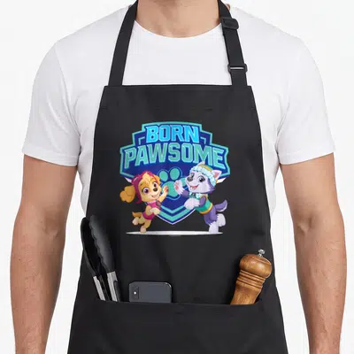 Pawsome isle of skye apron - BORN PAWSOME text in blue shield with two cartoon puppies in front — shown on a black cotton canvas apron against a white background
