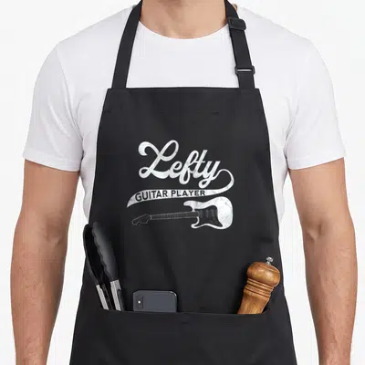 Guitarist left apron - White distressed Lefty GUITAR PLAYER text above a horizontal electric guitar on black fabric — shown on a black cotton canvas apron against a white background