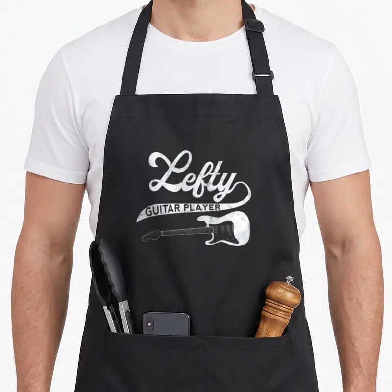 Guitarist left apron - White distressed Lefty GUITAR PLAYER text above a horizontal electric guitar on black fabric — shown on a black cotton canvas apron against a white background