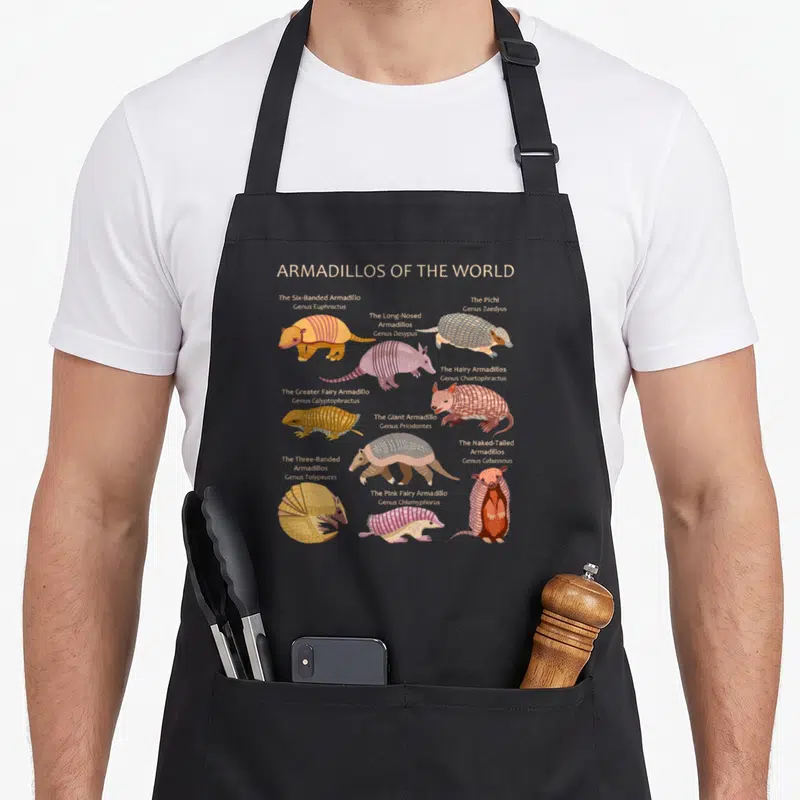 Specy armadillo apron - Ten cartoon armadillos with scientific names and pink orange grey brown shells — shown on a black cotton canvas apron against a white background