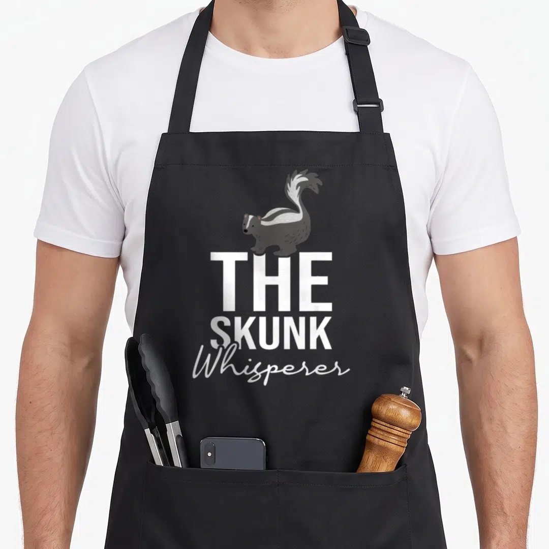 Skunk animal apron - Cartoon skunk standing on white THE SKUNK Whisperer text — shown on a black cotton canvas apron against a white background