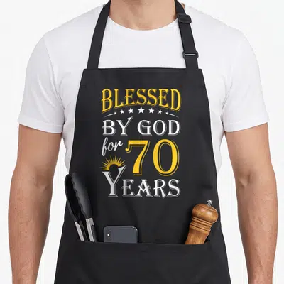 God 70th birthday apron - Stacked yellow and white text reading BLESSED BY GOD for 70 YEARS with stars and sunburst — shown on a black cotton canvas apron against a white background