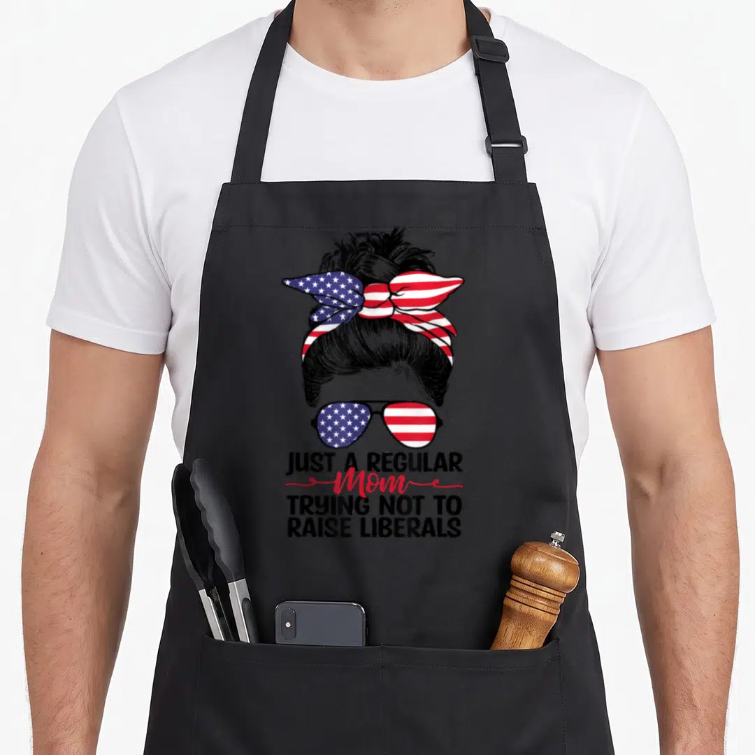 Trying mom apron - Woman with messy bun and flag sunglasses above text trying not to raise liberals — shown on a black cotton canvas apron against a white background