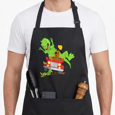 Dino decade apron - Green dinosaur chasing red jeep with brown dog and flying yellow hat in cartoon style — shown on a black cotton canvas apron against a white background
