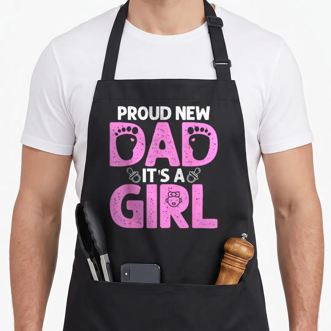 Funny proud father day apron - PROUD NEW DAD IT'S A GIRL text with pink footprints and baby face illustration — shown on a black cotton canvas apron against a white background