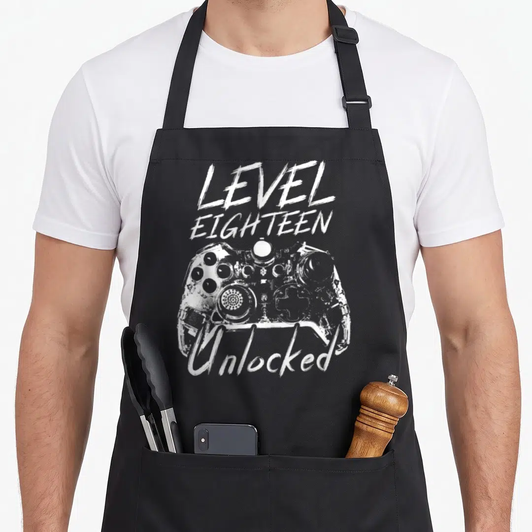 18th birthday party apron - White distressed game controller with LEVEL EIGHTEEN Unlocked text overlay — shown on a black cotton canvas apron against a white background