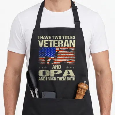 Title veteran grandpa apron - Distressed USA flag with soldier silhouettes and text I HAVE TWO TITLES VETERAN AND OPA — shown on a black cotton canvas apron against a white background
