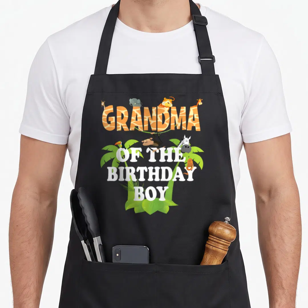 Zoo grandma apron - Cartoon animals and palm leaves surrounding GRANDMA OF THE BIRTHDAY BOY text in orange and white — shown on a black cotton canvas apron against a white background