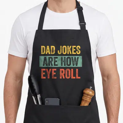 Joke father day apron - DAD JOKES ARE HOW EYE ROLL text in yellow, grey, and orange distressed font — shown on a black cotton canvas apron against a white background