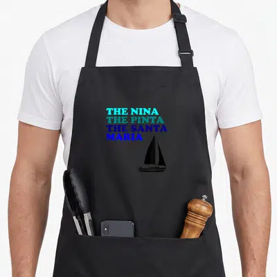 Nina santa apron - Stacked text reading The Nina The Pinta The Santa Maria beside a black sailboat silhouette — shown on a black cotton canvas apron against a white background