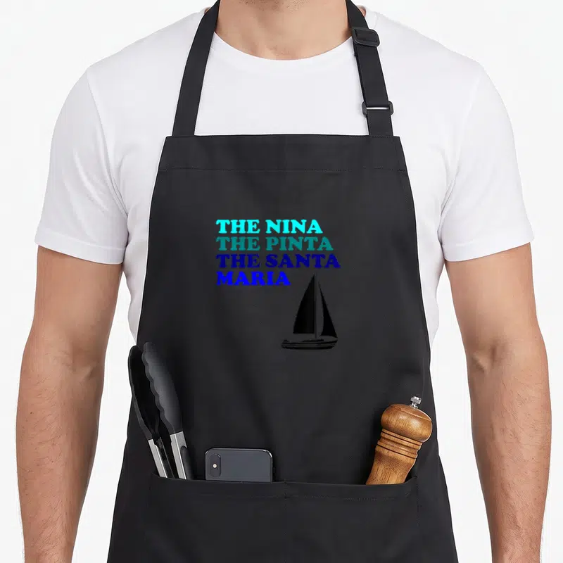 Nina santa apron - Stacked text reading The Nina The Pinta The Santa Maria beside a black sailboat silhouette — shown on a black cotton canvas apron against a white background