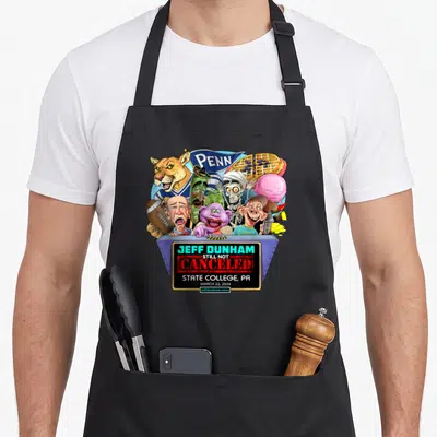Jeff venue apron - Caricatures on blue podium with Still Not Canceled text and Penn banner — shown on a black cotton canvas apron against a white background