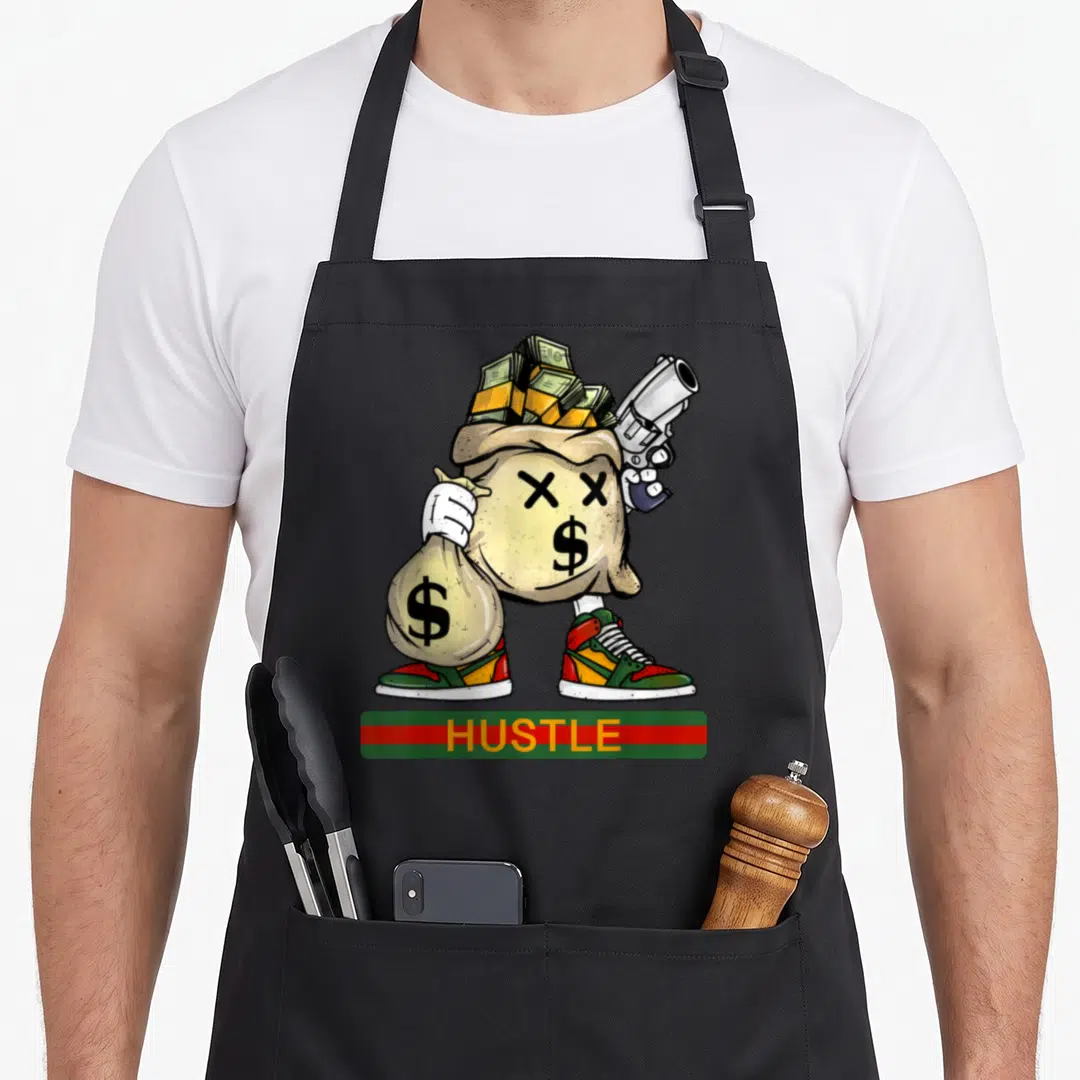 Stack money apron - Cartoon money bag with X eyes holding gun and cash, HUSTLE text in red green bar — shown on a black cotton canvas apron against a white background