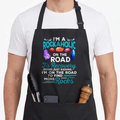 Rockaholic colorful aprons - I'M A ROCKAHOLIC text with five colorful rocks and crystals stacked vertically — shown on a black cotton canvas apron against a white background