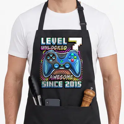 Level unlocked 7th video game apron - Blue game controller with LEVEL 7 UNLOCKED and AWESOME SINCE 2015 text in cartoon — shown on a black cotton canvas apron against a white background