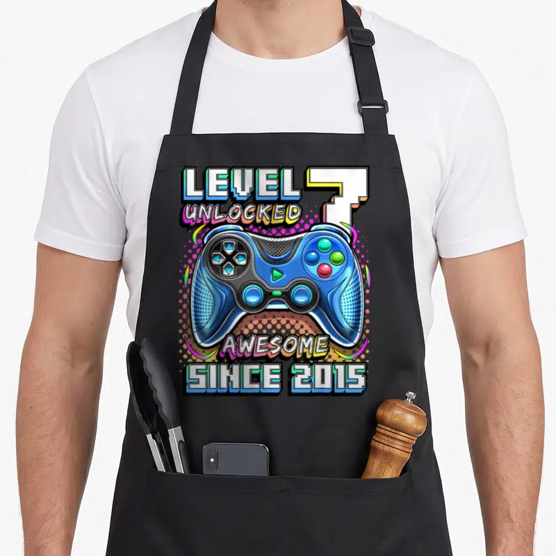 Level unlocked 7th video game apron - Blue game controller with LEVEL 7 UNLOCKED and AWESOME SINCE 2015 text in cartoon — shown on a black cotton canvas apron against a white background