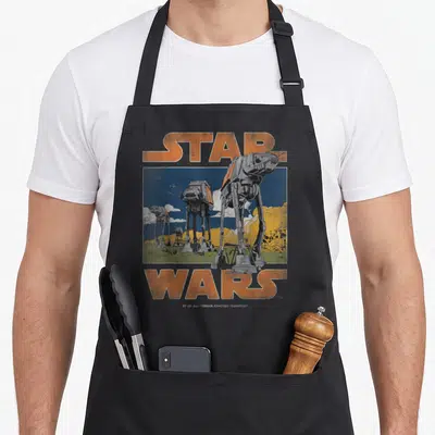Cute walker apron - Retro Star Wars AT-AT walker illustration with orange text and blue sky background — shown on a black cotton canvas apron against a white background