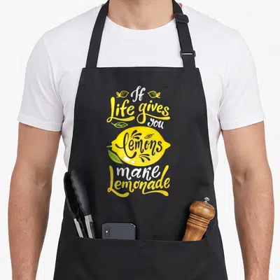 Give lemon lemonade apron - Stacked yellow and white text reading If life gives you lemons make lemonade around a large — shown on a black cotton canvas apron against a white background