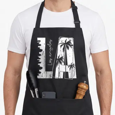 Funny los angeles apron - White LA block letters with grey skyline and palm trees, vertical script text on black — shown on a black cotton canvas apron against a white background