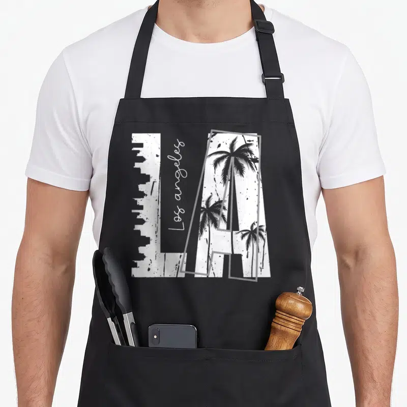 Funny los angeles apron - White LA block letters with grey skyline and palm trees, vertical script text on black — shown on a black cotton canvas apron against a white background