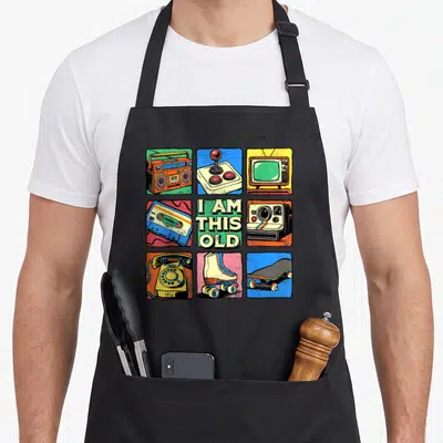 Nostalgia 80s apron - Grid of boombox, joystick, TV, cassette, camera, phone, skates, skateboard with text I AM THIS OLD — shown on a black cotton canvas apron against a white background