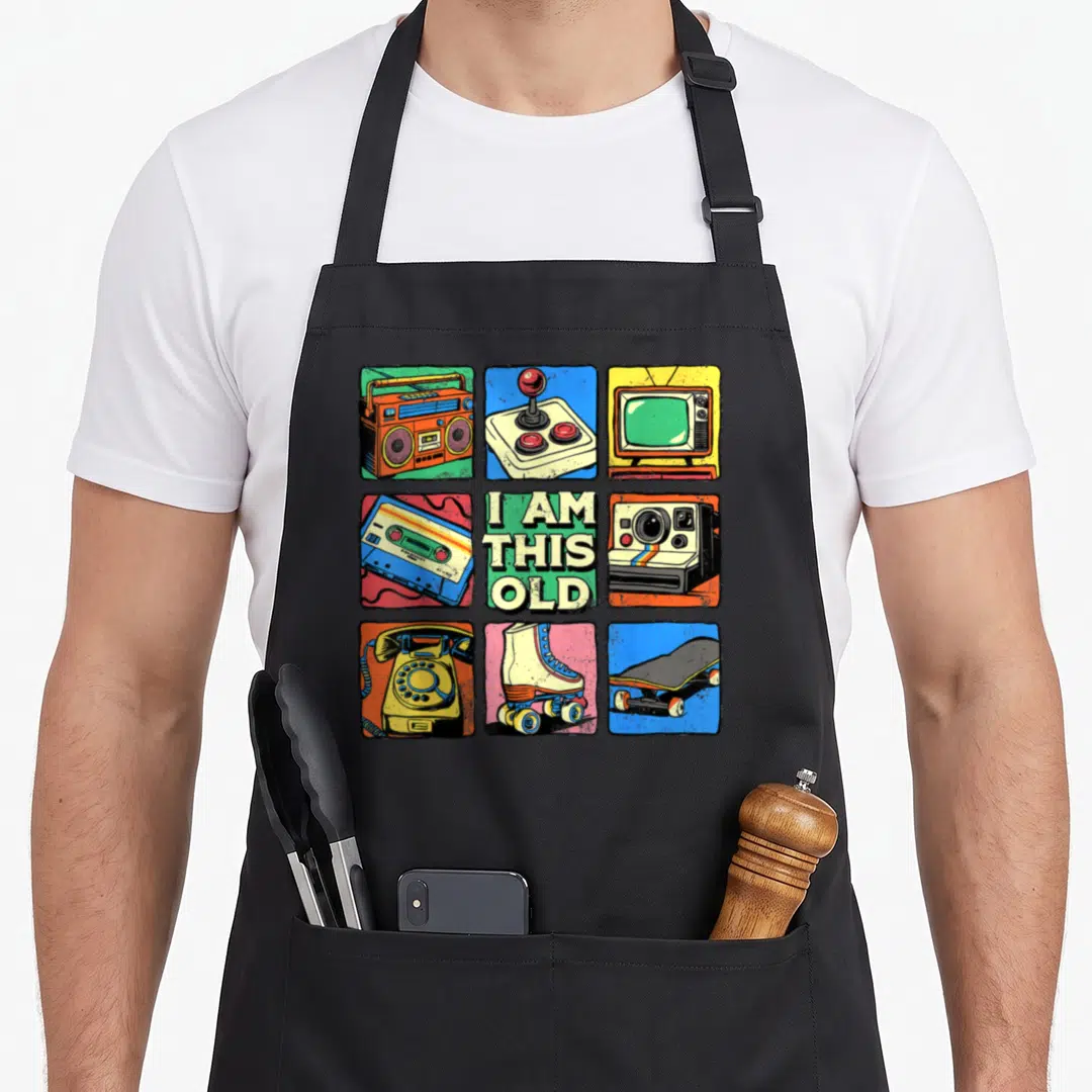 Nostalgia 80s apron - Grid of boombox, joystick, TV, cassette, camera, phone, skates, skateboard with text I AM THIS OLD — shown on a black cotton canvas apron against a white background