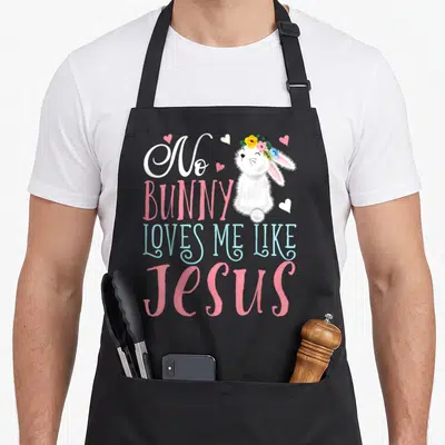 Jesus bunny apron - Cartoon white bunny with flower crown next to stacked pink and blue text reading No Bunny Loves Me — shown on a black cotton canvas apron against a white background