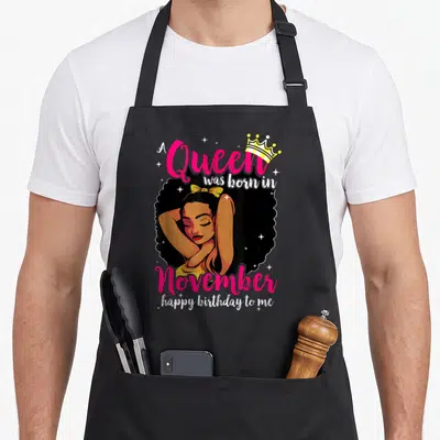 Afro diva queen birthday apron - Cartoon woman with crown bow and A Queen was born in November happy birthday to me text — shown on a black cotton canvas apron against a white background